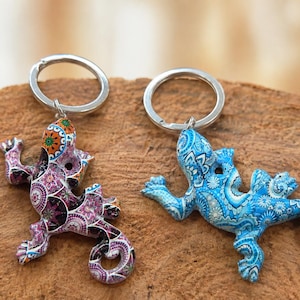 Op de afbeelding: Twee hagedisvormige sleutelhangers met metalen ringen. De ene hagedis heeft een paars en zwart mandala-patroon, terwijl de andere een blauw en wit ingewikkeld bloemendesign toont. Beide figuren hebben gekrulde staarten en gedetailleerde texturen.
