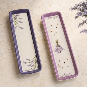 May include: Two rectangular ceramic serving trays with raised edges. The left tray has a deep purple rim and lavender sprig illustrations. The right tray features a lighter purple rim, a central lavender bouquet, and small butterfly motifs.