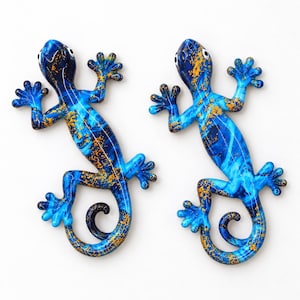 Könnte beinhalten: Zwei Gecko-Figuren mit einem leuchtend blauen und dunkelblauen Marmormuster und verstreuten goldfarbenen Sprenkeln. Jeder Gecko hat einen geringelten Schwanz, vier gespreizte Füße mit sichtbaren Zehen und weiße Augen mit schwarzen Pupillen.