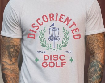 May include: A white t-shirt with a red and blue graphic design that says "DISCORIENTED" with a disc golf basket and the words "SINCE 1975 DISC GOLF".