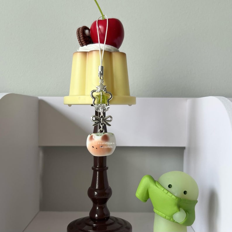 May include: A decorative charm featuring a yellow pudding with a cherry and cookie on top. A silver charm with a green gem and a small apple-shaped charm with text hang below. A small green figure is in the background.