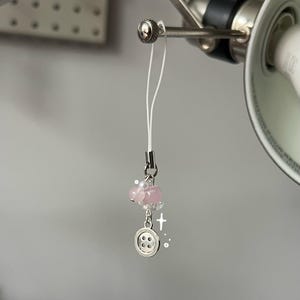 May include: A delicate phone charm featuring a white cord, pink beads, and a silver button charm. The charm includes a small cross and a pearl accent, creating a whimsical and feminine accessory.