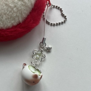 May include: A white and pink ceramic charm with a green design, attached to a silver chain and a red fluffy accessory. The charm features a flower and star design. The accessory is a small, decorative item.