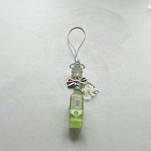 May include: A green miniature bottle charm with Japanese text, a silver bow, and clear beads. The charm has a silver loop for attachment. The bottle is filled with a light green liquid, and the charm is on a white background.