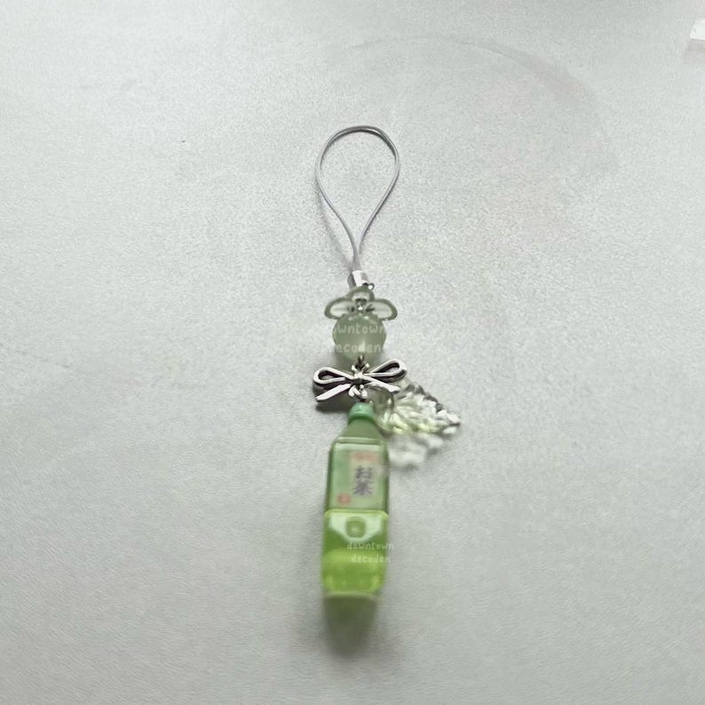 May include: A green miniature bottle charm with Japanese text, a silver bow, and clear beads. The charm has a silver loop for attachment. The bottle is filled with a light green liquid, and the charm is on a white background.