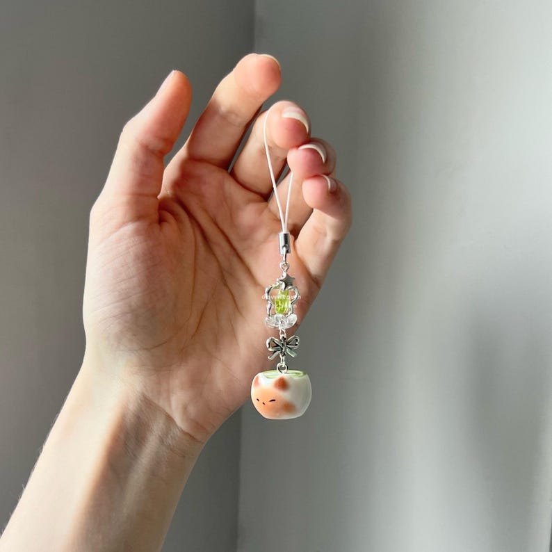 May include: A silver-toned charm with a white cord, featuring a small, round, ceramic object with orange and white spots. The charm also includes a crown, a green gem, and a bow. The charm is held in a hand.