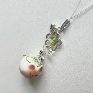 May include: A cute, white and brown ceramic charm with a matcha-like design, featuring a small face. The charm is attached to a silver-colored chain with a flower-shaped accent and a star. A white string is attached to the top.