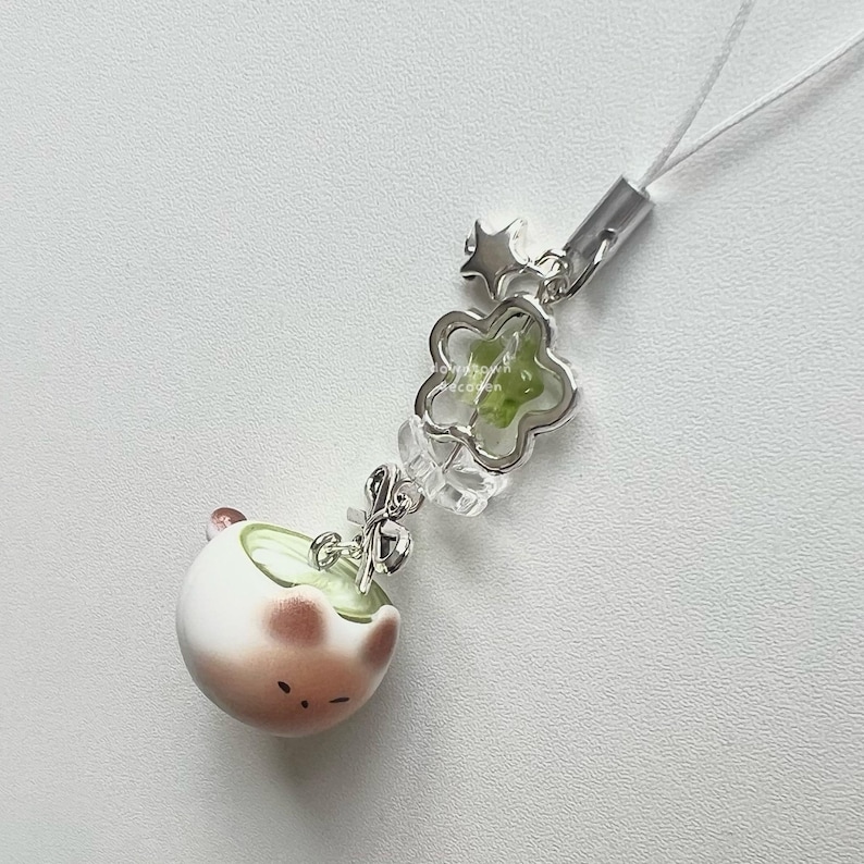 May include: A cute, white and brown ceramic charm with a matcha-like design, featuring a small face. The charm is attached to a silver-colored chain with a flower-shaped accent and a star. A white string is attached to the top.