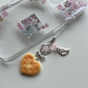 May include: Clear phone case with a decorative charm. The charm features a heart-shaped biscuit, a silver bow, a flower-shaped charm with text, a pink flower, and a silver wing. The case has various cute stickers inside.