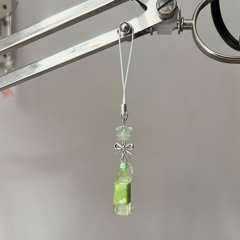 May include: A miniature green bottle charm with a white string, silver bow, and light green beads. The bottle has a label and is suspended from a silver clasp. The charm is hanging from a silver metal object.