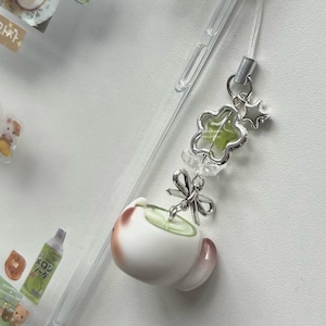 May include: A clear phone case with a decorative charm. The charm features a white and brown teacup with green liquid, a silver bow, a star, and a flower-shaped charm with green accents. A white cord is attached to the phone case.