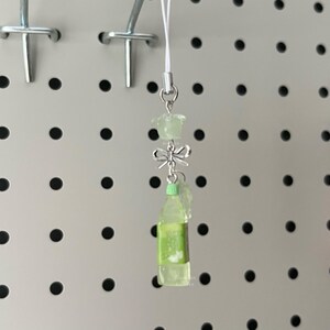 May include: A clear, lime green charm with a miniature bottle design. The charm features a small bow and a flower-shaped bead. The charm is hanging from a white string, and is attached to a silver metal clasp.