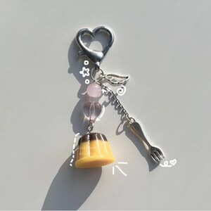 May include: A silver charm keychain with a heart-shaped charm, a small angel wing charm, and a pink and clear glass bead charm. The keychain also features a small, brown and yellow pudding charm with a silver spoon charm.
