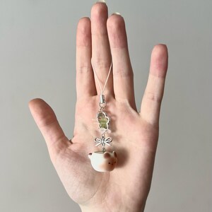May include: A small, cream-colored ceramic charm in the shape of a teacup with a bear face. The charm is attached to a silver-colored metal flower and bow, with clear beads and a white cord. The charm is held in a hand.
