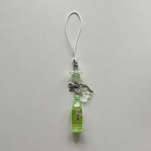 May include: A green and white phone charm featuring a miniature bottle of tea with Japanese text. The charm includes a clear, leaf-shaped embellishment, a small bow, and a flower-shaped bead. A white cord is attached for hanging.