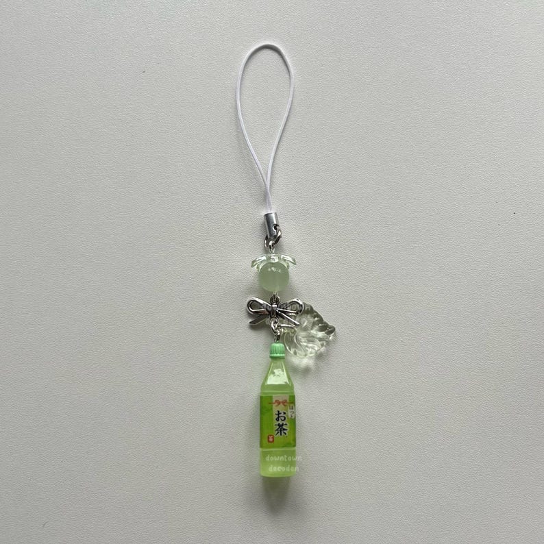 May include: A green and white phone charm featuring a miniature bottle of tea with Japanese text. The charm includes a clear, leaf-shaped embellishment, a small bow, and a flower-shaped bead. A white cord is attached for hanging.