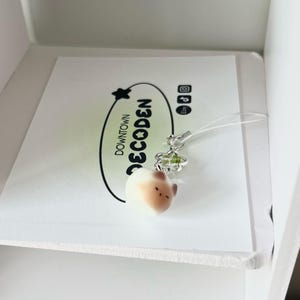 May include: A small, cream-colored charm in the shape of a rounded animal head with brown accents. It hangs from a silver-colored floral charm and a clear string. The charm rests on a white card with the text "DOWNTOWN DECODEN" in black.