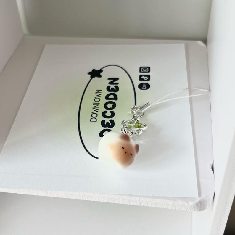 May include: A small, cream-colored charm in the shape of a rounded animal head with brown accents. It hangs from a silver-colored floral charm and a clear string. The charm rests on a white card with the text "DOWNTOWN DECODEN" in black.