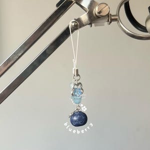 May include: A blue and white phone charm with a blueberry charm and a silver chain. The charm is hanging from a white string.