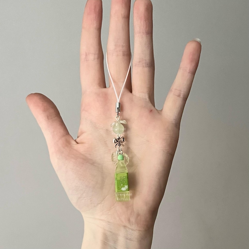 May include: A clear, green-toned charm with a white cord. The charm features a small, green bottle design, adorned with a silver bow and translucent flower-shaped beads. The charm is suspended from a white cord with a silver clasp.