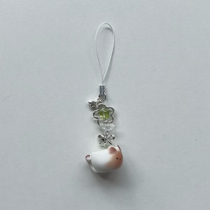 May include: A white and brown cat charm with a silver-colored bow, flower, and star accents. The charm is attached to a clear string. The cat charm is in the shape of a teacup.