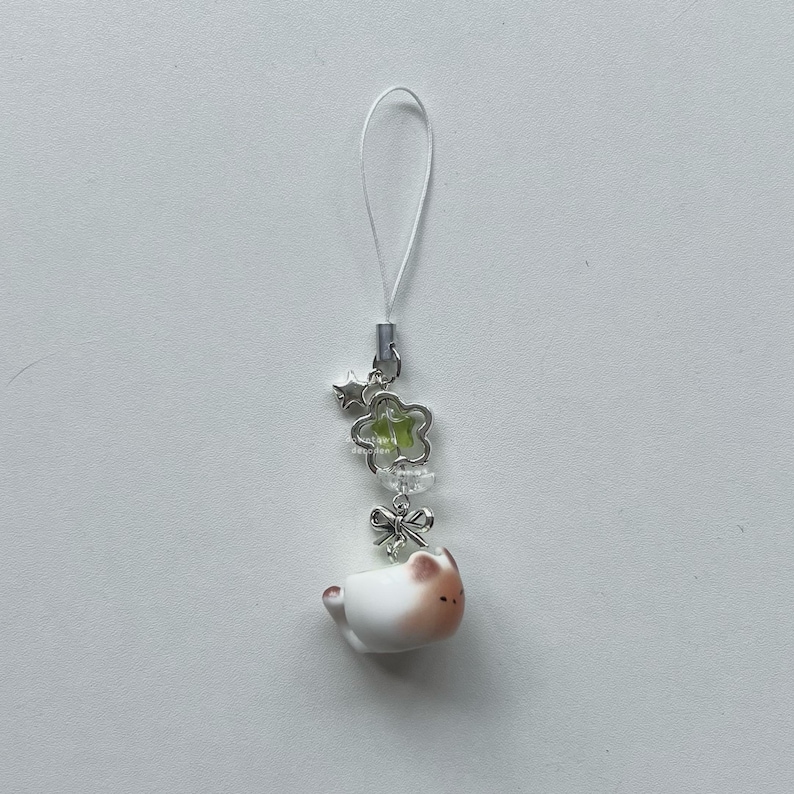 May include: A white and brown cat charm with a silver-colored bow, flower, and star accents. The charm is attached to a clear string. The cat charm is in the shape of a teacup.
