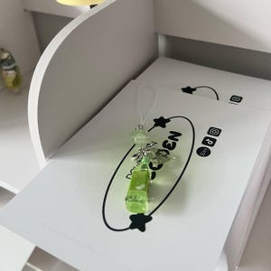 May include: A green and clear resin bottle charm with a silver bow and bead accent, attached to a white string. The charm is resting on a stack of white cards with the text "Golden" and social media icons.