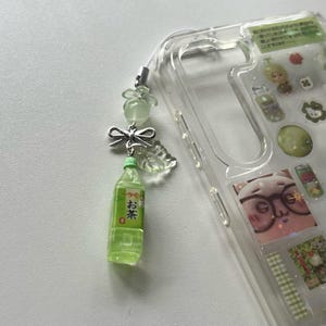 May include: A clear phone case with decorative stickers and a green tea bottle charm. The charm includes a small green bottle with Japanese text, a silver bow, and translucent green beads. The case is on a white surface.