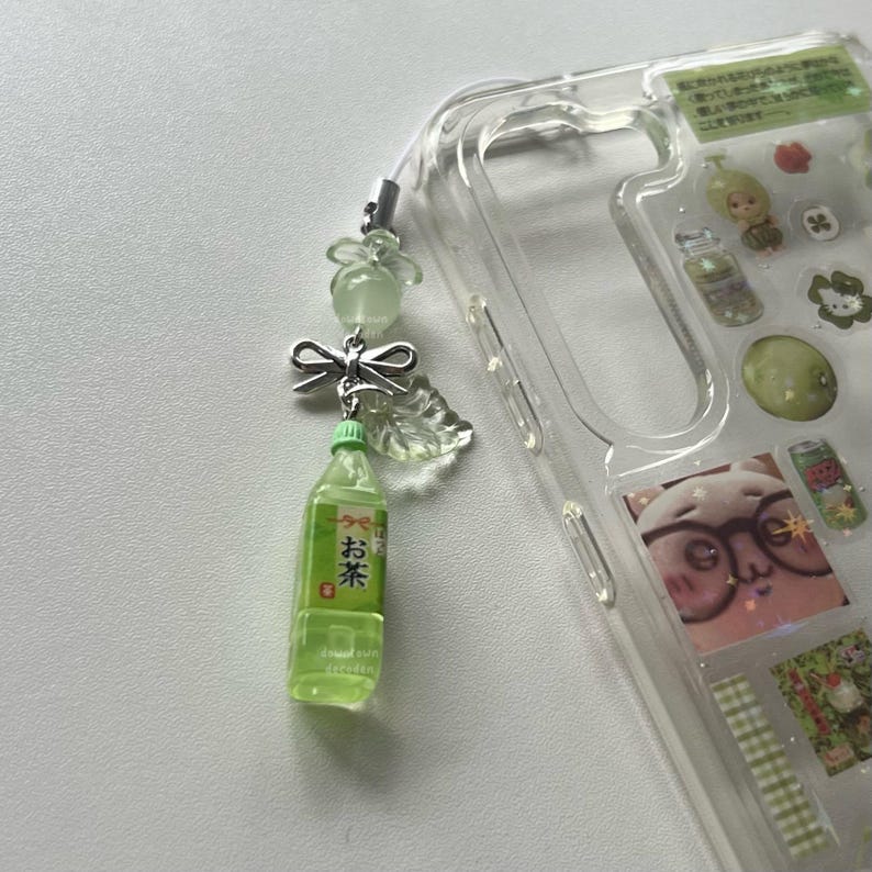 May include: A clear phone case with decorative stickers and a green tea bottle charm. The charm includes a small green bottle with Japanese text, a silver bow, and translucent green beads. The case is on a white surface.