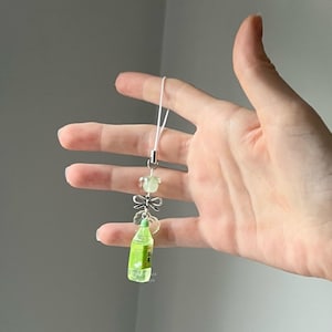May include: A clear, green bottle charm with Japanese text, accented with a silver bow, clear beads, and a light green stone. The charm is attached to a white cord, creating a delicate accessory.