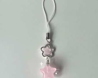 Puede incluir: Un charm de estrella rosa con un charm de flor plateada unido a una correa de teléfono blanca.