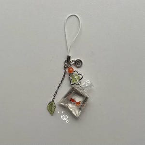 May include: A clear, plastic charm featuring a small goldfish in a bag. The charm is adorned with a chain, green leaf, flower, orange beads, and a white loop. The goldfish is orange and white. The charm is on a white background.