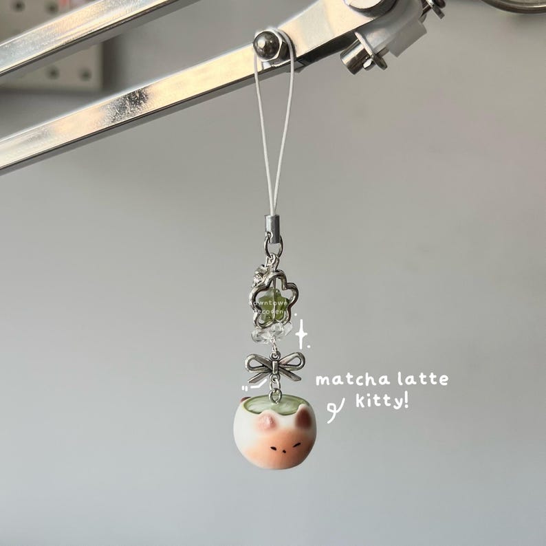 May include: A decorative charm featuring a small, round, light-colored kitty face with a green matcha latte design. The charm is suspended from a silver-colored chain with a bow and flower accents. Text on the side reads "matcha latte kitty!"