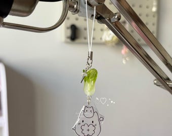 May include: A clear acrylic charm with a green bok choy bead and a cat and clover design. The charm is attached to a silver-colored metal clasp with a white string. The charm is hanging from a silver-colored metal stand.