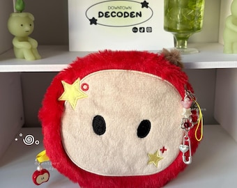 May include: A round, red plush pouch with a beige center panel featuring two black embroidered eyes. The pouch is decorated with two yellow star appliques and a dangling charm set including a pink flower, a red bead, and a silver heart pin.