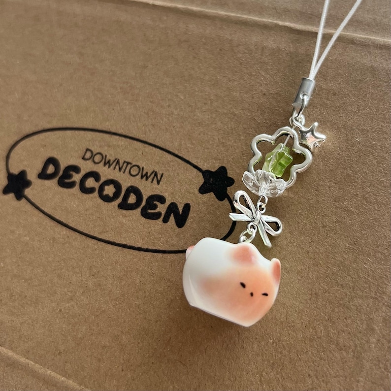 May include: A decorative phone charm featuring a small, white and pink piglet figurine. The charm includes a silver-toned floral design with a green gem and a clear crystal, along with the text "Downtown Decoden".