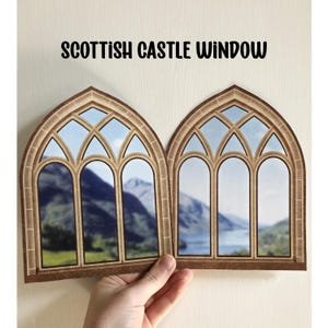 Puede incluir: Una ventana de papel marrón hecha a mano con vista a un lago y montañas. La ventana está diseñada para parecerse a una ventana de castillo escocés con ventanas arqueadas y mampostería.