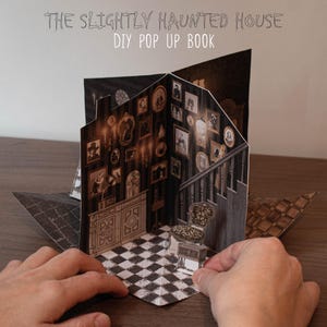 May include: A DIY pop-up book titled "The Slightly Haunted House" featuring a detailed interior scene. The book's interior depicts a room with framed portraits, a staircase, and a checkered floor. The book is open, revealing the pop-up design.