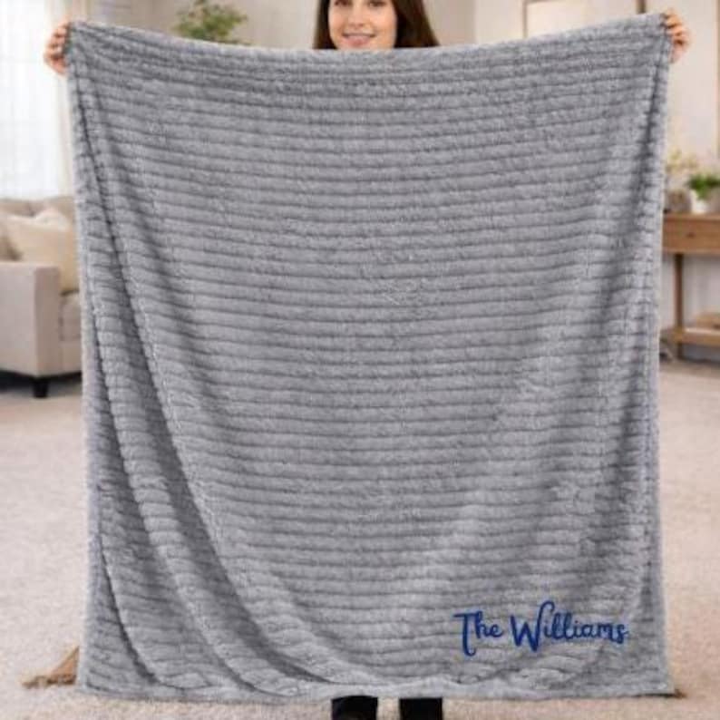 May include: A gray, plush blanket with a ribbed texture is held up. The blanket has a soft, furry appearance and is embroidered with the name "The Williams" in a cursive font. The blanket is displayed indoors.