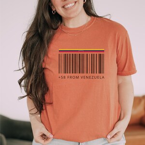 May include: Burnt orange t-shirt featuring a barcode design with the colors of the Venezuelan flag at the top. The text "+58 FROM VENEZUELA" is printed below the barcode. The shirt is a short-sleeved, casual style.