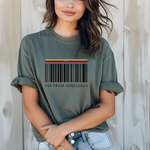 May include: Olive green t-shirt with a barcode design and the text "+58 FROM VENEZUELA" in black. The barcode has a colorful stripe at the top, representing the Venezuelan flag. The shirt is a casual, short-sleeved style.