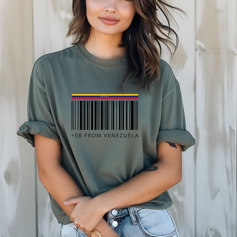 May include: Olive green t-shirt with a barcode design and the text "+58 FROM VENEZUELA" in black. The barcode has a colorful stripe at the top, representing the Venezuelan flag. The shirt is a casual, short-sleeved style.