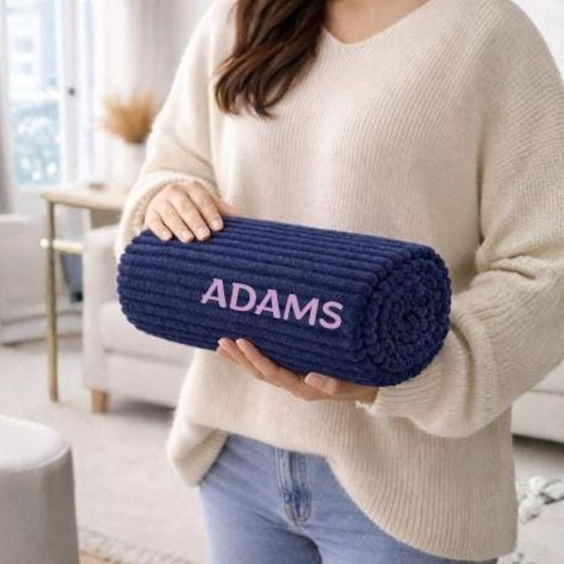May include: A navy blue, cylindrical pillow with the name "ADAMS" in pink lettering. The pillow has a ribbed texture and is held by a person wearing a cream-colored sweater and blue jeans.