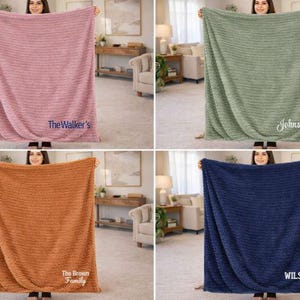 May include: Four plush blankets in different colors: dusty rose, sage green, burnt orange, and navy blue. Each blanket has a textured pattern and a personalized name or family name embroidered on it. The blankets are displayed in a well-lit room.