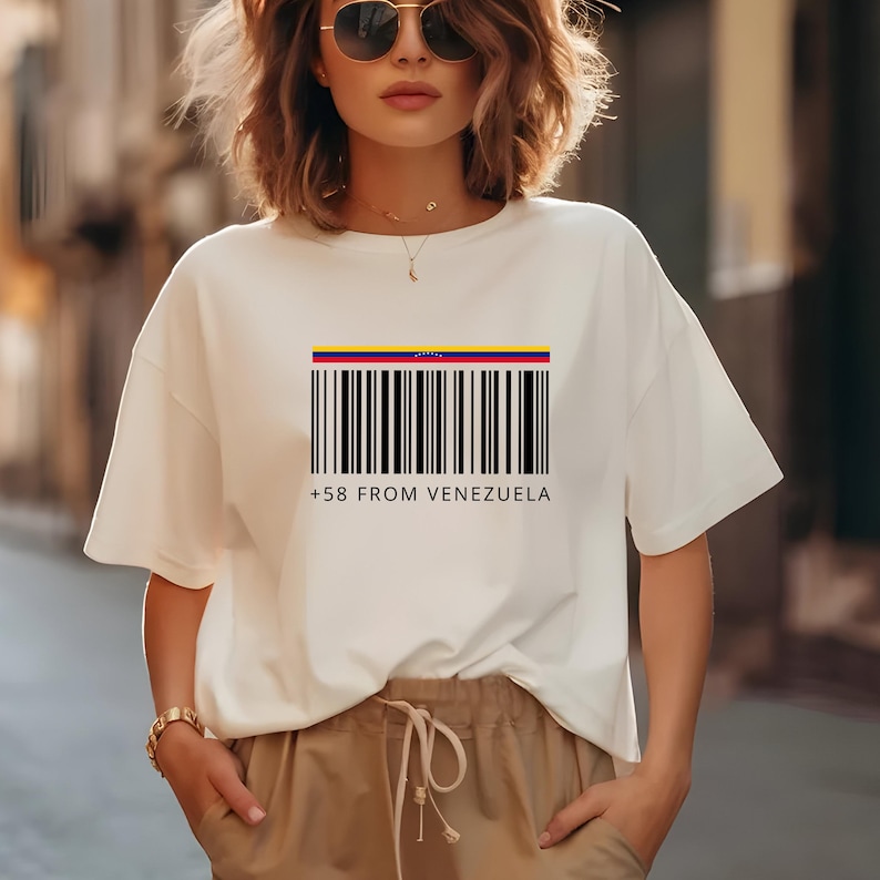 May include: White t-shirt featuring a barcode design with the colors of the Venezuelan flag at the top. Below the barcode is the text "+58 FROM VENEZUELA" in a black font. The shirt has short sleeves and a relaxed fit.