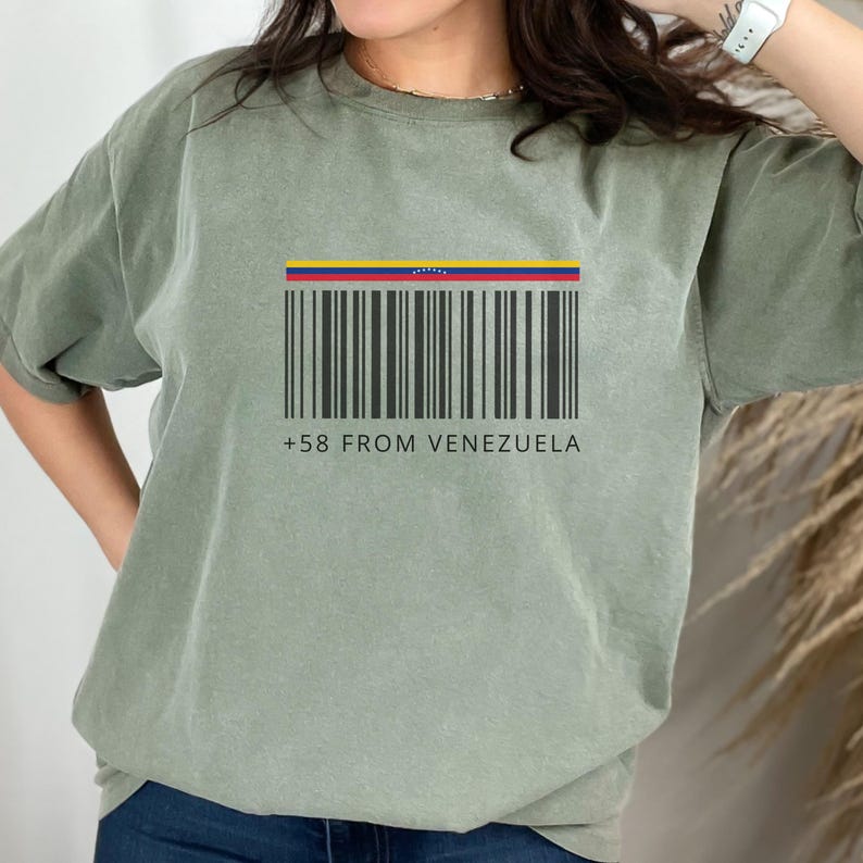 May include: Sage green t-shirt featuring a barcode design with the colors of the Venezuelan flag at the top. Below the barcode is the text "+58 FROM VENEZUELA" in a simple, sans-serif font.