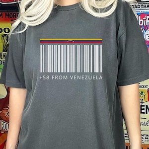 May include: Dark gray t-shirt featuring a white barcode design with the colors of the Venezuelan flag at the top. The text "+58 FROM VENEZUELA" is printed below the barcode. The shirt is a casual wear item.