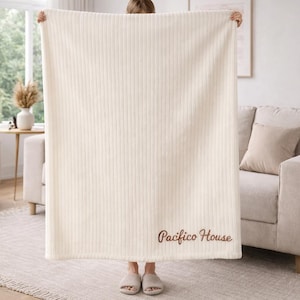 May include: Cream-colored, vertically ribbed blanket with the embroidered text "Pacifico House" in a brown script. The blanket is held up, revealing a glimpse of a living room with a sofa, rug, and window.