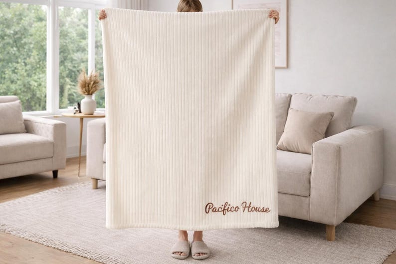 May include: Cream-colored, vertically ribbed blanket with the embroidered text "Pacifico House" in a brown script. The blanket is held up, revealing a glimpse of a living room with a sofa, rug, and window.