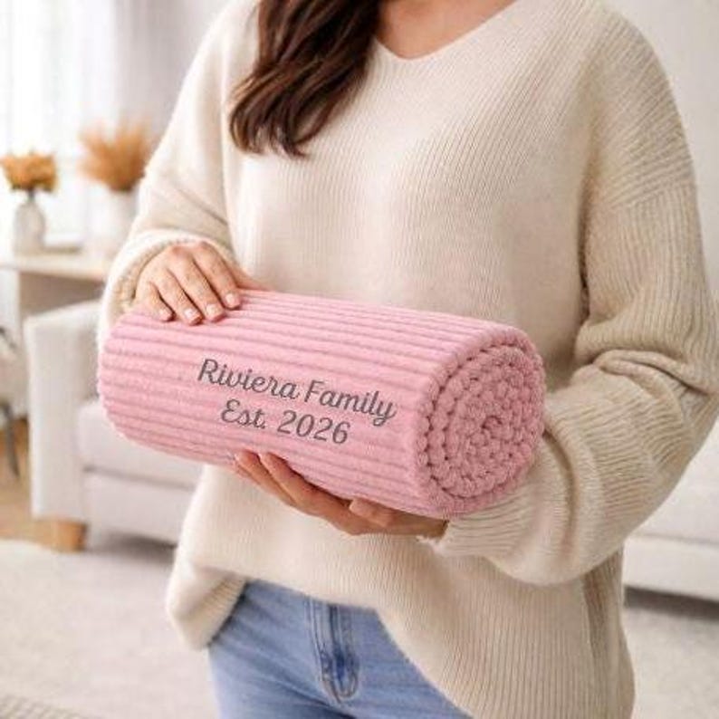 May include: A rolled-up, light pink blanket with a ribbed texture. The blanket has the text "Riviera Family Est. 2026" embroidered on it. The blanket is held by a person wearing a cream-colored sweater and blue jeans.